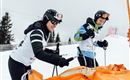 Zwei Skifahrer bereiten sich auf einen Wettkampf vor. Sie tragen Schutzkleidung und stehen auf einer verschneiten Piste.