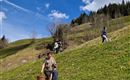 A green meadow with some trees and a blue sky. A person is carrying a basket, while a child stands nearby.