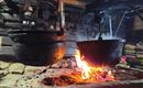 Ein rustikaler Holzofen mit zwei hängenden Kochtöpfen über einem offenen Feuer. Die Flammen leuchten warm und schaffen eine gemütliche Atmosphäre.