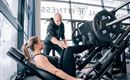 Eine Frau trainiert im Fitnessstudio, während sie Unterstützung von einem Trainer erhält. Die Geräte und die große Fensterfront sorgen für ein modernes Ambiente.