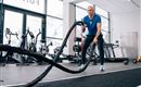 Ein Mann trainiert im Fitnessstudio mit einem Battle Rope. Der Raum ist modern und gut ausgestattet mit verschiedenen Fitnessgeräten.