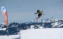 A skier jumps over a snow hill. In the background, there are snow-covered mountains and a clear sky.
