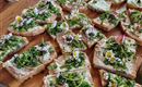 A wooden board with various slices of bread topped with herbs and edible flowers. The presentation is fresh and inviting.