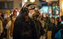 Ein kostümierter Mann mit einem furchterregenden Kopf und Hörnern steht in einer festlichen Menschenmenge. Es schneit, und im Hintergrund sind Lichter und weitere kostümierte Personen zu sehen.