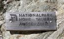 Ein Holzschild mit der Aufschrift "NATIONALPARK HOHE TAUERN AUSSENZONE". Es befindet sich an einem felsigen Hintergrund.