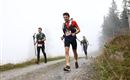 Drei Läufer bewegen sich auf einem schmalen Weg durch neblige Landschaft. Im Hintergrund sind einige Bäume zu sehen.