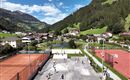 Ein malerischer Blick auf ein Sportgelände mit Tennisplätzen und einem Freizeitraum. Im Hintergrund erstrecken sich grüne Hügel und ein malerisches Dorf.