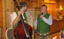 Zwei Männer in traditioneller Tracht stehen in einem gemütlichen Raum. Einer spielt Kontrabass, während der andere ein Mikrofon hält und spricht.