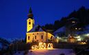 Eine bezaubernde Kirche im Schnee, hell erleuchtet gegen den Nachtblau-Himmel. Im Vordergrund sind Weihnachtsdekorationen mit Figuren zu sehen.