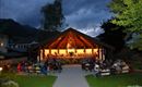 A beautiful venue with wooden architecture, surrounded by trees and mountains. Many people are sitting on benches and enjoying an outdoor event at dusk.