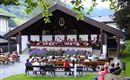 An orchestra is playing outdoors under a pavilion. Many people are sitting on benches and enjoying the music.