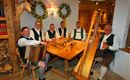 Eine Gruppe von sechs Männern in traditionellen Trachten sitzt an einem Holztisch. Sie spielen Instrumente und genießen Bier in einer gemütlichen, rustikalen Umgebung.