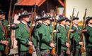 Eine Gruppe von Männern in traditionellen grünen Uniformen steht aufrecht und hält Gewehre in der Hand. Sie scheinen an einer Zeremonie oder Parade teilzunehmen.