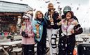 Sieben Frauen feiern den Gewinn von Pokalen in einem festlichen Moment auf einer Skihütte. Im Hintergrund fallen Konfetti und es schneit leicht.