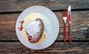 Ein köstliches Dessert auf einem weißen Teller mit Vanillesauce und Puderzucker. Daneben liegen eine Gabel und ein Messer auf einem rustikalen Holztisch.