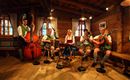 Eine Gruppe von fünf Musikern in traditioneller bayerischer Tracht spielt in einem rustikalen Raum. Sie verwenden verschiedene Instrumente, darunter ein Akkordeon und eine Gitarre.