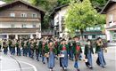 Eine festliche Parade mit Menschen in traditionellen Trachten, die musikalische Instrumente tragen. Im Hintergrund sind typische Holzgebäude und Bäume zu sehen.