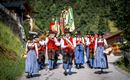Eine Gruppe von Menschen in traditionellen Trachten marschiert eine Straße entlang. Sie tragen instruments und halten Fahnen in der Hand, umgeben von malerischer Natur.