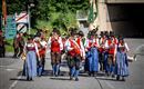 Eine Gruppe von Musikern marschiert auf einer Straße, gekleidet in traditionelle Trachten. Sie spielen verschiedene Instrumente und tragen Fähnchen.