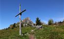 Ein schöner Bergweg mit einem Holzkreuz und einer Sitzgelegenheit. Im Hintergrund sind majestätische Berge und ein klarer blauer Himmel zu sehen.