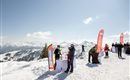 A sunny winter landscape with snow and mountains in the background. People are enjoying an event with standing tables and brand flags at the summit.