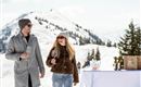 A couple enjoys a sunny day in the mountains, holding wine glasses in their hands. In the background, there are snow-covered mountains and a table with wine bottles.