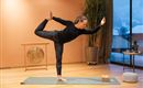 A woman is performing a yoga exercise in a modern room. The atmosphere is calm and inviting, with large windows and plants in the background.