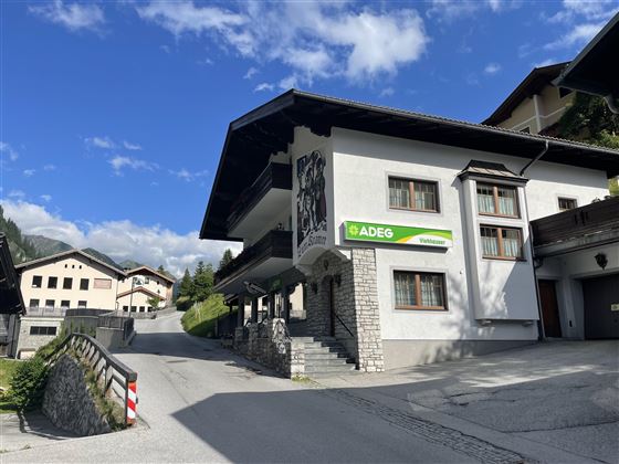 Ein Gebäude mit traditioneller Architektur und einem ADEG-Schild. Die Straße führt sanft bergauf, umgeben von malerischen Bergen.