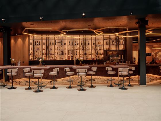 Eine moderne Bar mit einer eleganten Theke und stilvollen Barhockern. Im Hintergrund sind Regale mit Getränken und warmes Licht zu sehen.