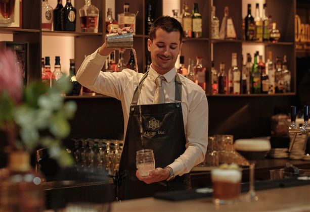 Ein Barkeeper in einem eleganten Raum mixt einen Drink. Im Hintergrund sind Regale mit verschiedenen Spirituosen sichtbar.