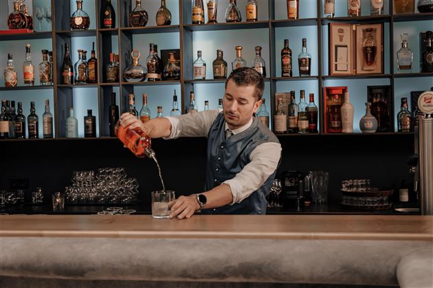 Ein Barkeeper gießt einen Cocktail in ein Glas. Im Hintergrund sind viele Flaschen Alkohol ordentlich aufgestellt.