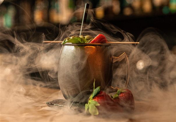 Ein Cocktail in einem silbernen Becher, umgeben von dampfender Atmosphäre. Frische Erdbeeren und Kräuter ergänzen das Getränk.