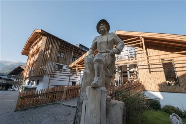 A wooden sculpture of a man sits on a tree trunk in front of modern, wooden-clad buildings. The sky is clear and the surroundings are surrounded by mountains.