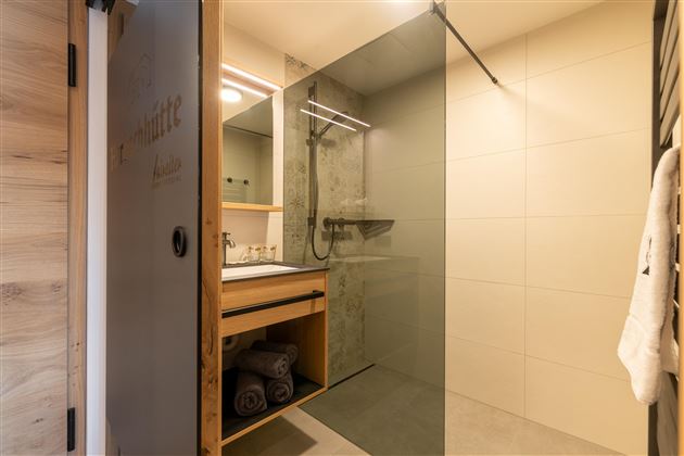 A modern bathroom with a glass shower and a wooden shelf. The decor is simple and elegant.