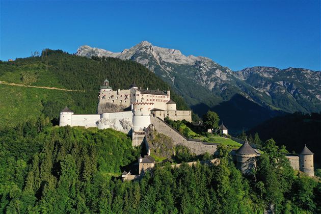 A majestic castle on a green hill, surrounded by mountains. The clear blue sky above the castle completes the picturesque landscape.