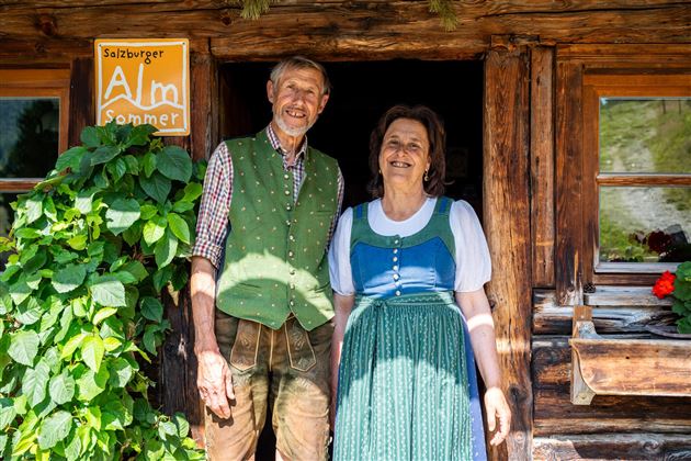 Ein älteres Paar steht vor einer rustikalen Holzhütte. Beide tragen traditionelle Kleidung und lächeln freundlich in die Kamera.