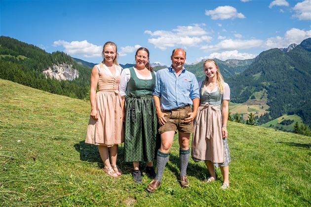 Eine Gruppe von vier Personen im traditionellen bayerischen Trachten steht auf einer Wiese mit Bergen im Hintergrund. Der Himmel ist blau mit wenigen Wolken.