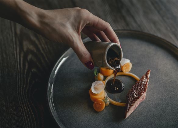 A hand pours a dark sauce over an artfully arranged dish on a gray plate. The dish includes colorful garnishes like pumpkin and fresh herbs.