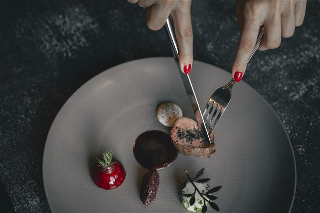 An elegant dish on a plate with various components. A hand is cutting into the food with a knife and fork.