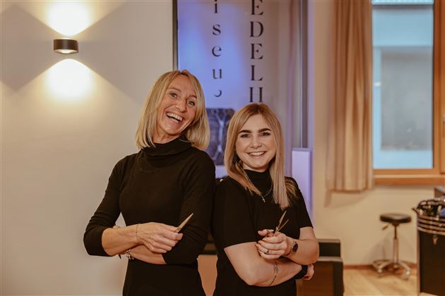 Zwei lächelnde Friseurinnen stehen nebeneinander in einem modernen Salon. Sie tragen schwarze Kleidung und halten Friseurzubehör in der Hand.