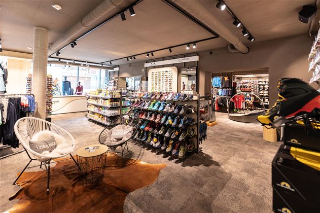 Ein modernes Sportgeschäft mit einer Vielzahl von Schuhen und Kleidung. Es gibt gemütliche Sitzgelegenheiten und eine helle, einladende Atmosphäre.