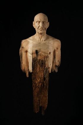 A sculptural representation of a human torso made of wood. The head is richly detailed, while the lower part ends in irregular wooden fragments.