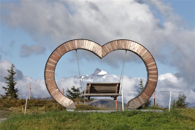 A wooden sculpture in the shape of a heart with a bench inside. In the background, tall mountains and a cloudy sky can be seen.