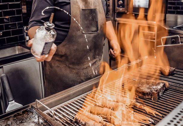 Ein Koch gart gegrillte Meeresfrüchte auf einem heißen Grill, während Flammen aufsteigen. Der Koch sprüht eine Flüssigkeit auf das Grillgut, um das Flammenspiel zu verstärken.