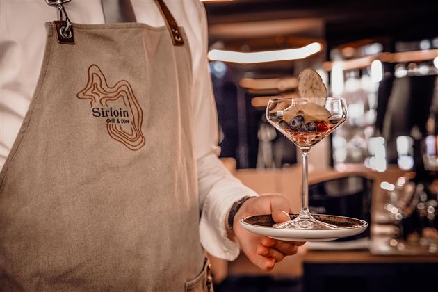 Ein Kellner in einem Schürze hält ein Dessertglas mit Beeren und Eis. Im Hintergrund ist eine stilvolle Restaurantatmosphäre zu sehen.