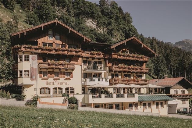 Ein charmantes Hotel im alpinen Stil, umgeben von grünen Hügeln. Die Balkone sind mit Blumen geschmückt und bieten einen einladenden Anblick.