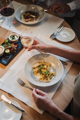 A table with elegant food and drinks. A person holds a glass of white wine and enjoys a bowl of pasta.