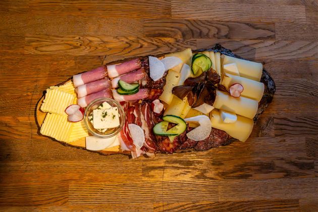 Eine Platte mit verschiedenen Käsesorten, Wurst und frischem Gemüse. Darauf befindet sich auch eine Schale mit Aufstrich.