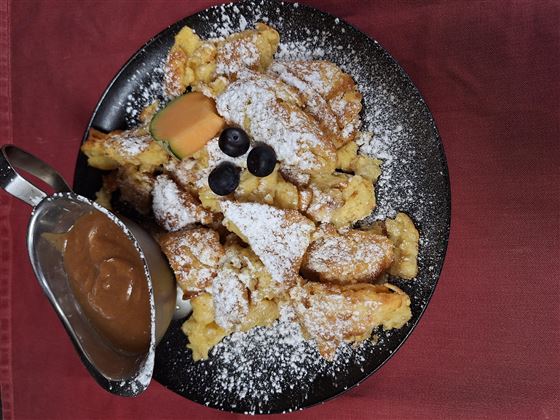 Ein Teller mit süßem Kaiserschmarrn, bestreut mit Puderzucker und frischen Früchten. Daneben befindet sich eine Sauce in einer kleinen Kanne.
