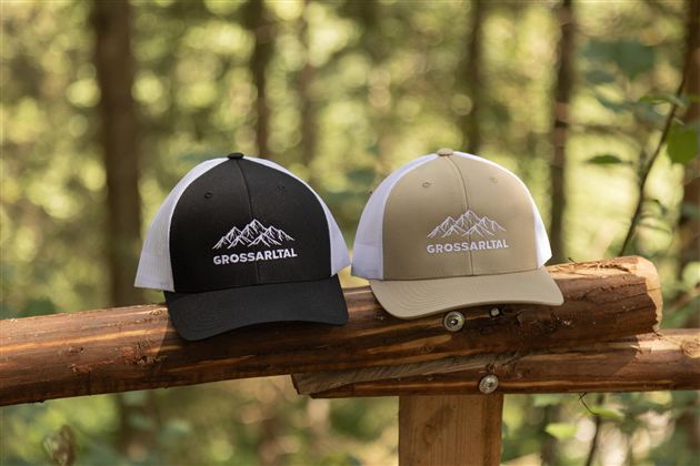 Two caps are standing on a wooden railing in the forest. One cap is black and the other is khaki, both featuring a mountain motif and the inscription "GREAT".